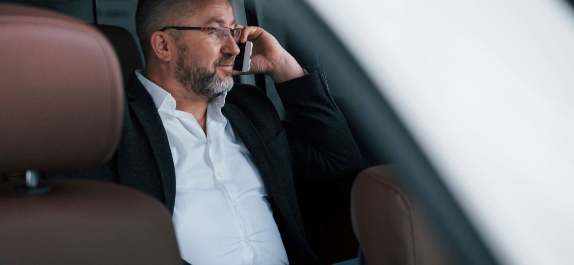 Calm and good mood. Having business call while sitting at the back of modern luxury car. Senior man in glasses and official wear