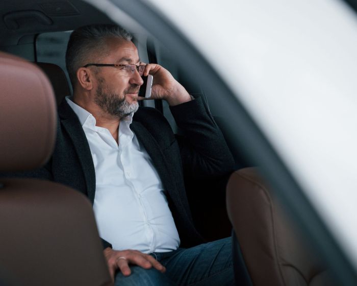 Calm and good mood. Having business call while sitting at the back of modern luxury car. Senior man in glasses and official wear